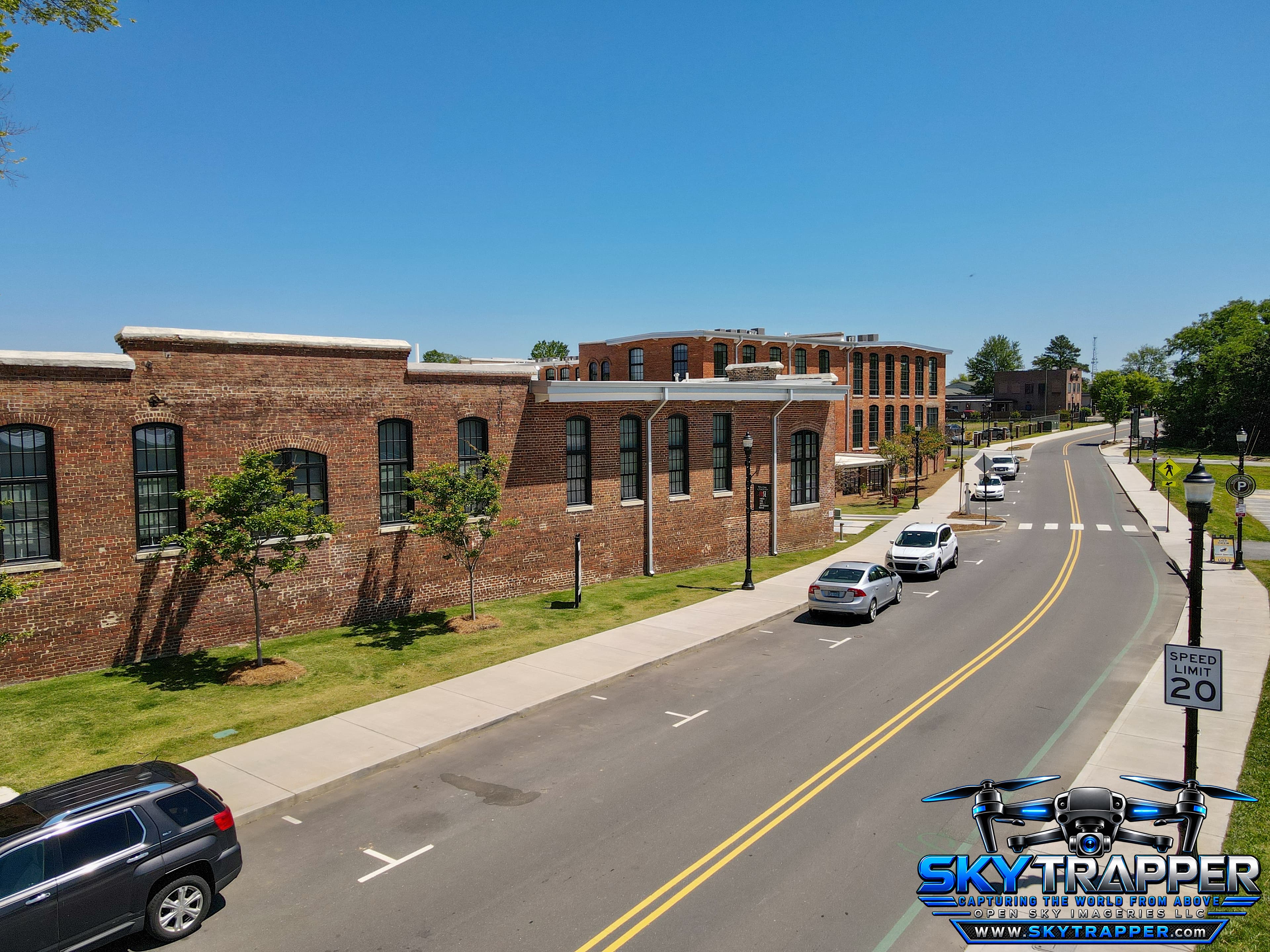 Aerial drone photograph of restored red-brick industrial buildings along a two-lane street, featuring sidewalks, parked vehicles, landscaping, and surrounding architecture.