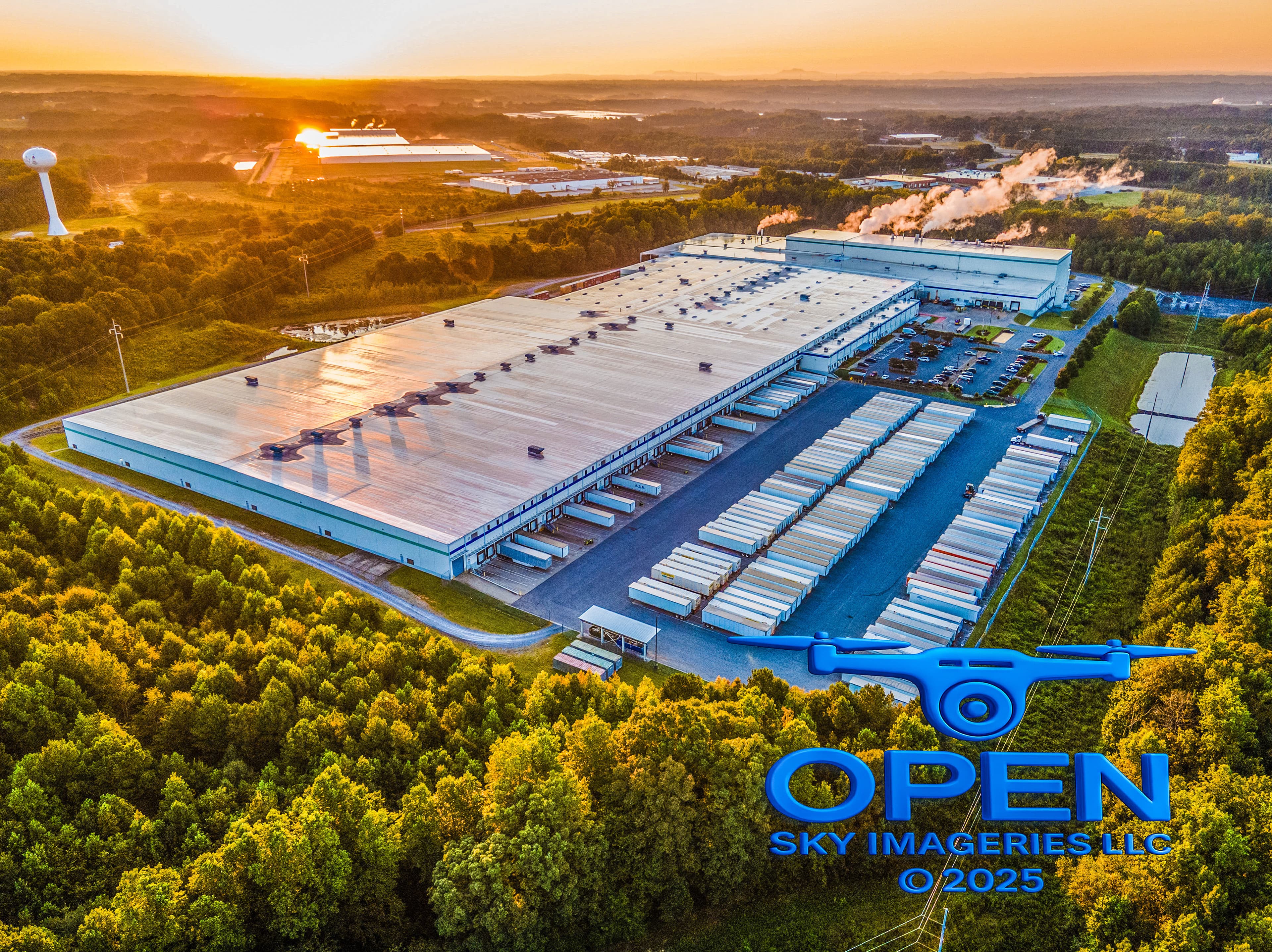 Golden-hour aerial photograph of a large industrial warehouse and distribution complex with loading docks, rows of parked trailers, parking areas, surrounding woodland, and nearby industrial buildings.