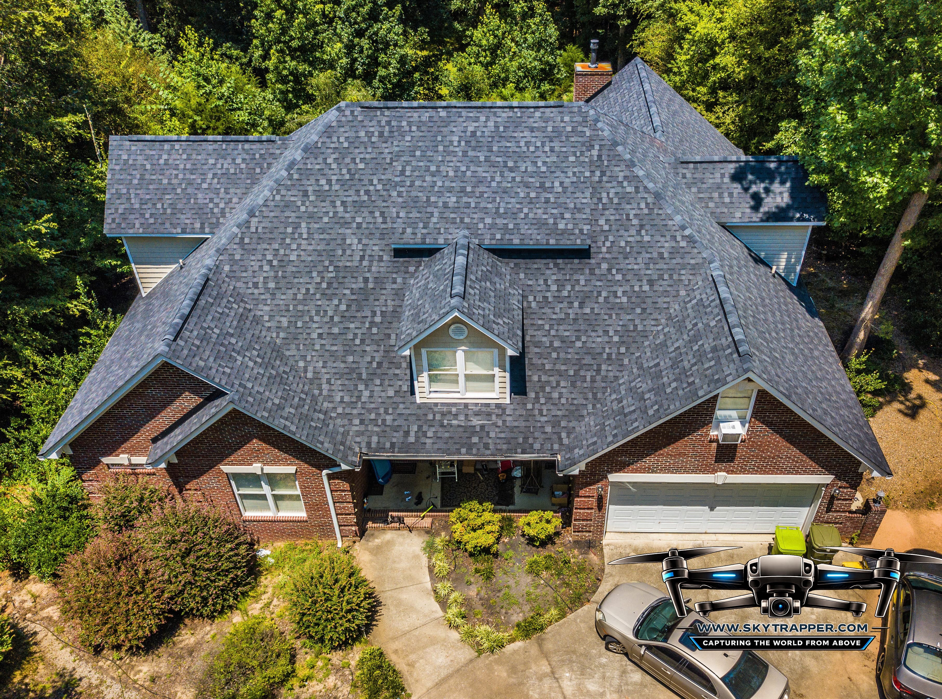 Aerial drone photograph of a brick residential home featuring a large gray architectural shingle roof, multiple roof slopes, a central dormer window, chimney, attached two-car garage, landscaped front yard, and surrounding trees.