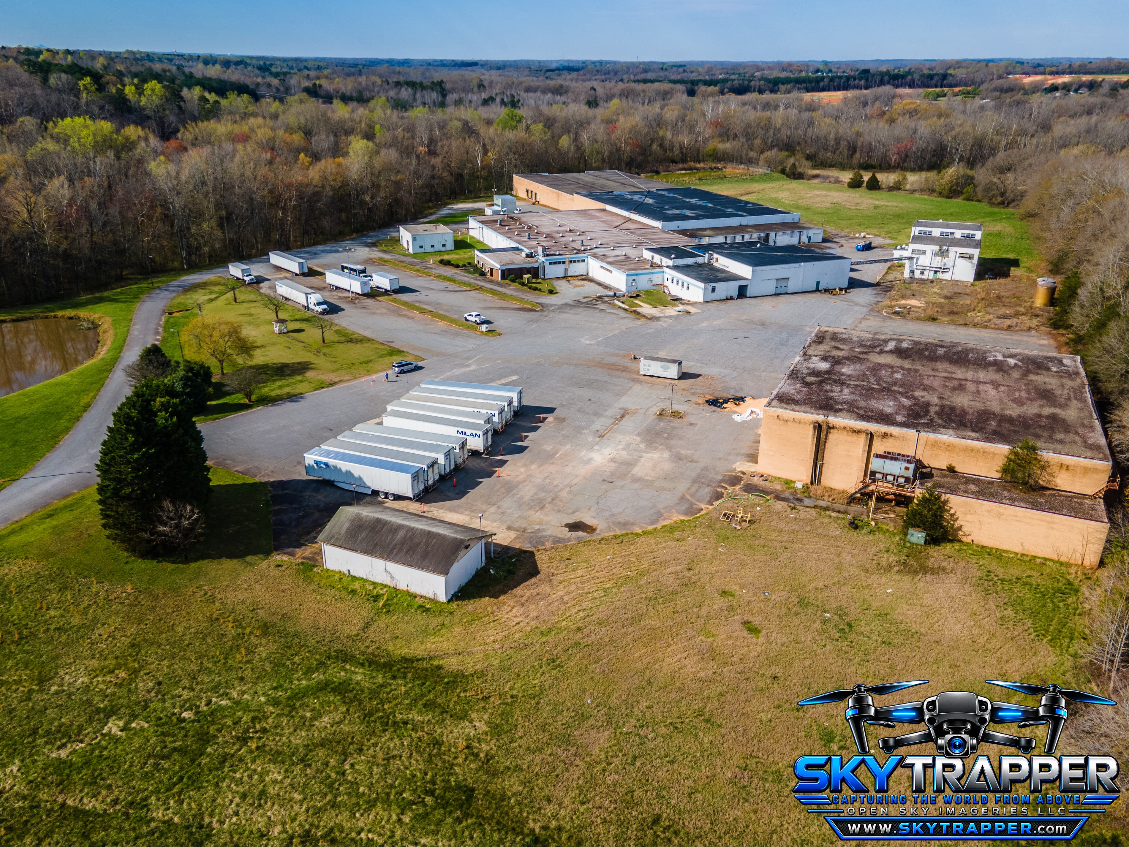 Aerial drone view of a large industrial facility with multiple warehouse buildings, loading areas, parked semi-trailers, expansive paved lots, surrounding green space, and wooded land.